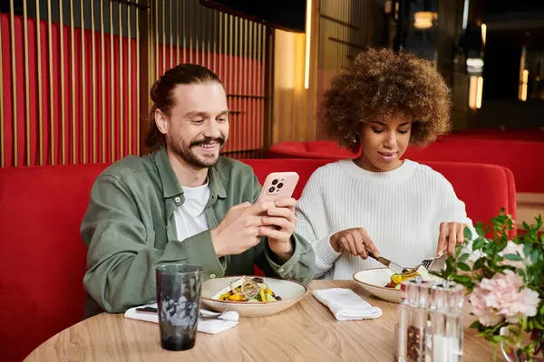Un uomo e una donna, assorti con un telefono cellulare e cibo a un tavolo da caffè. — Foto stock