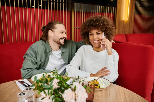 Una mujer afroamericana y un hombre profundamente involucrados en conversaciones telefónicas mientras están sentados en una mesa de café. - foto de stock