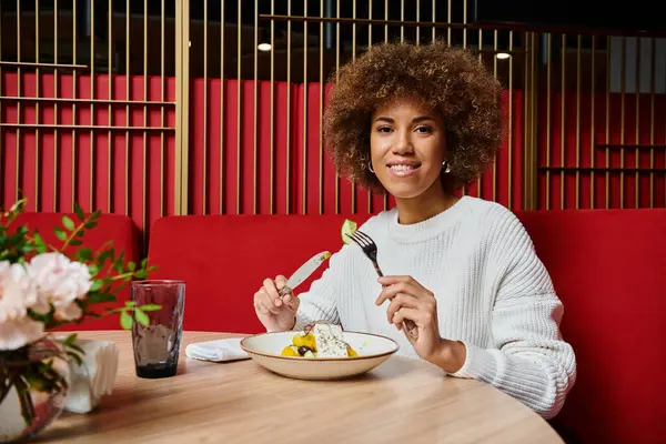 Uma mulher afro-americana senta-se à mesa, desfrutando de uma tigela de comida em um café moderno com um homem nas proximidades. — Fotografia de Stock
