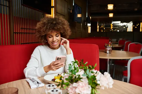 Une Afro-Américaine s'assoit à une table, absorbée par son téléphone portable, dans un café moderne. — Photo de stock