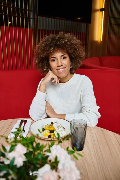 Uma mulher afro-americana elegante senta-se em uma mesa, desfrutando de um prato de comida deliciosa em um café moderno, enquanto profundamente no pensamento. — Fotografia de Stock