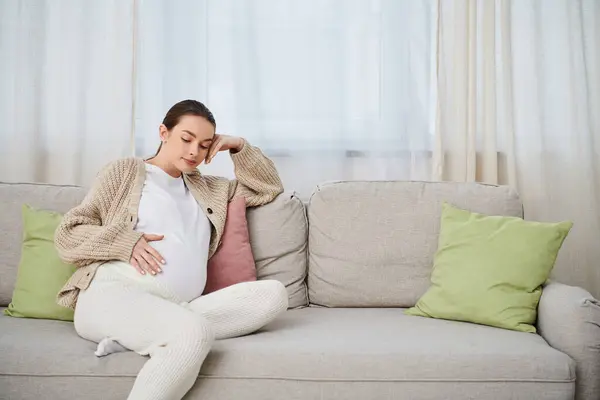Una hermosa mujer embarazada se sienta cómodamente en un sofá en una acogedora sala de estar, rodeada de calidez y anticipación. - foto de stock