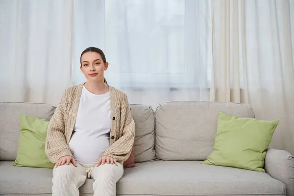 Eine schöne schwangere Frau sitzt auf einer Couch, ihre Silhouette wird vom weichen Licht eines Fensters beleuchtet. — Stockfoto