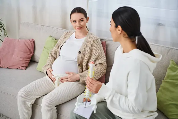 Вагітна жінка сидить на дивані, зайнявшись розмовою з іншою вагітною жінкою, ймовірно, її тренером під час батьківських курсів. — стокове фото