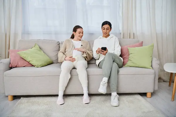 Un uomo e una donna incinta siedono su un divano, assorti in uno schermo del telefono cellulare, probabilmente condividono un momento di connessione.. — Foto stock