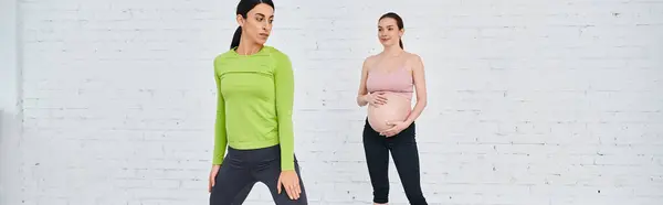 Una mujer está de pie junto a una mujer embarazada durante un curso de los padres, apoyándola y guiándola a través de ejercicios. - foto de stock