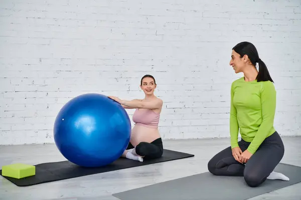 Две женщины, одна беременная, сидят на ковриках для йоги, занимаясь с большим мячом для упражнений под руководством тренера во время родительских курсов.. — стоковое фото