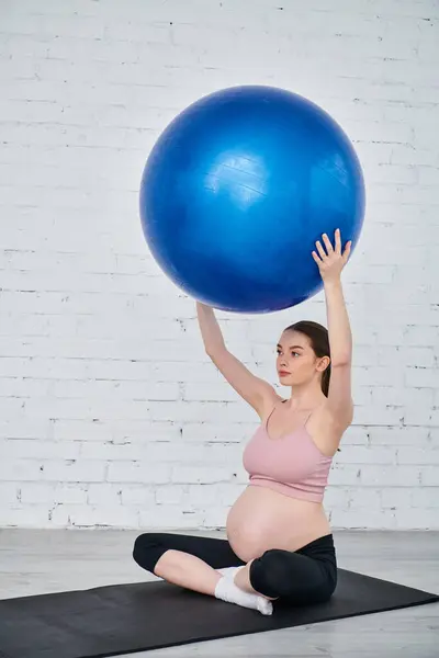 Pregnant woman in yoga pose on mat, holding blue ball during prenatal exercise session. — Stock Photo