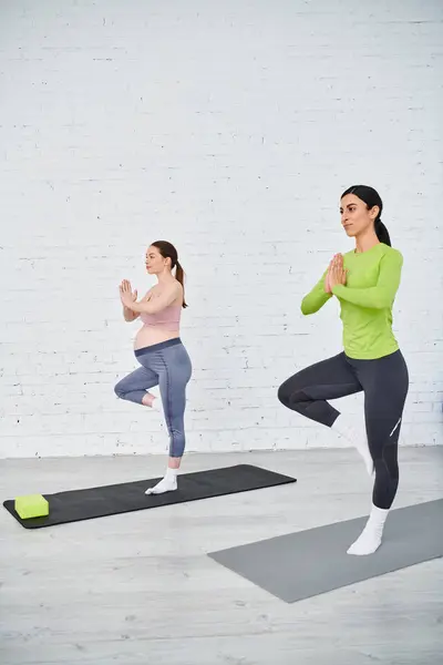 Two women, one pregnant, gracefully pose in yoga positions, finding peace and strength in front of a white brick wall. — Stock Photo