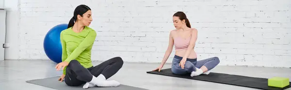 Dos mujeres, una embarazada, se sientan serenamente en colchonetas de yoga en un momento compartido de relajación y camaradería durante un curso para padres. - foto de stock