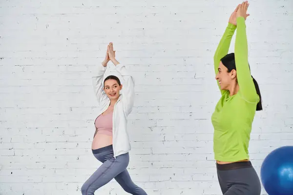 Una mujer embarazada se ejercita con su entrenador durante un curso de padres, apoyado por otra mujer de pie junto a ella. - foto de stock