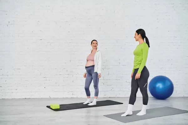 Two women, one pregnant, stand on yoga mats in a gym, engaged in a serene moment of mindfulness and movement during a parents course. — Stock Photo
