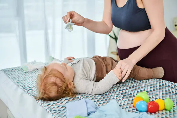 A young, beautiful mother tenderly holds soother near baby who is peacefully lying on a bed at home. — Stock Photo