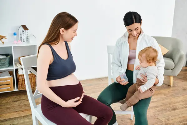Una mujer embarazada se sienta graciosamente en una silla, acunando a un bebé en su regazo con el apoyo de su entrenador en los cursos de los padres. - foto de stock