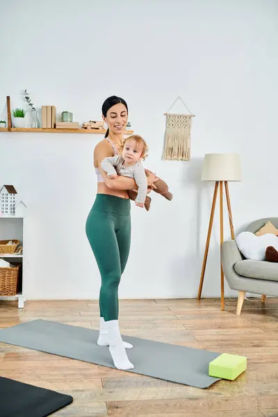 Eine junge Mutter steht auf einer Yogamatte und hält ihr Baby sanft in der Hand, angeleitet von einem Trainer bei Elternkursen. — Stockfoto