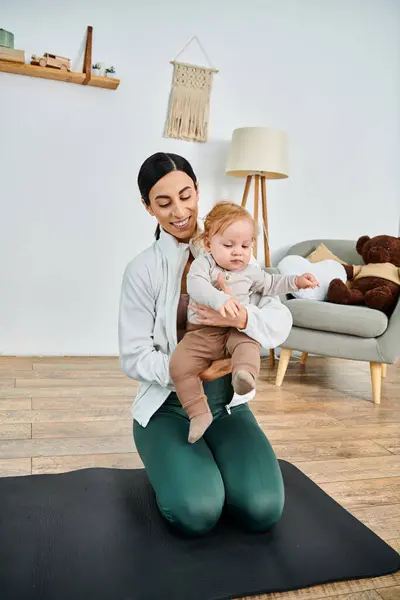 Una madre joven se sienta en una esterilla de yoga, sosteniendo pacíficamente a su bebé mientras recibe orientación de su entrenador durante un curso para padres.. - foto de stock