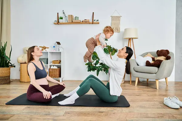 Una joven madre sentada tranquilamente en una esterilla de yoga con un niño sentado alegremente sobre sus hombros, guiada por un entrenador de apoyo. - foto de stock