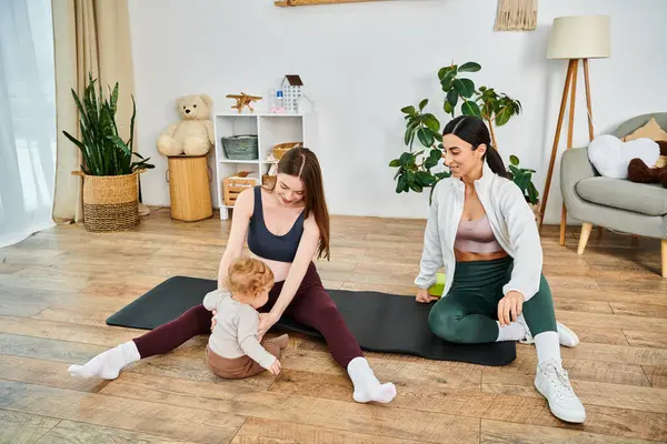 Dos mujeres, una madre joven y serena y su entrenador, guían a un bebé en una esterilla de yoga en un ambiente hogareño tranquilo. - foto de stock
