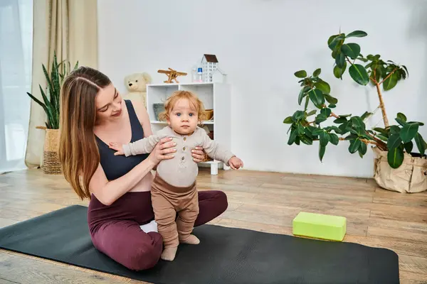 Una giovane madre siede su un tappetino da yoga che culla il suo bambino, guidata dal suo allenatore in un momento di pacifica connessione e cura. — Foto stock
