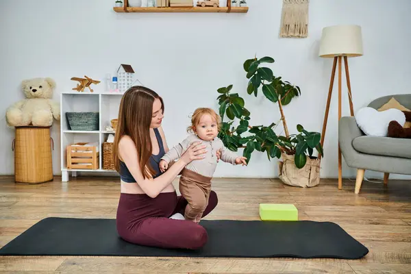 Una joven y hermosa madre sentada en una esterilla de yoga, acunando a su bebé con la guía de su entrenador durante un curso de padres. - foto de stock