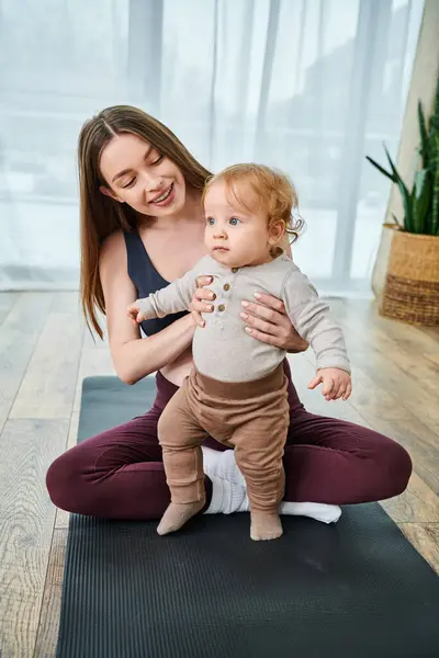 Una joven madre sostiene suavemente a su bebé en una esterilla de yoga, guiada por un entrenador en los cursos de los padres en la comodidad de su hogar. - foto de stock