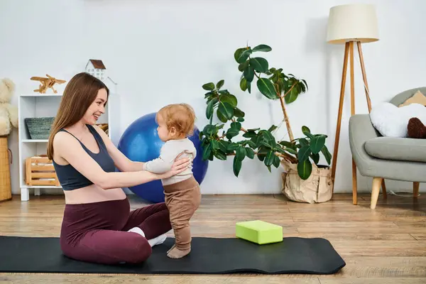 Una hermosa madre joven sentada en una esterilla de yoga, acunando a su bebé con la guía de un entrenador en los cursos de padres. - foto de stock