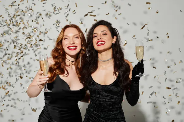 Two beautiful women in elegant attire clinking champagne flutes amid confetti. — Stock Photo