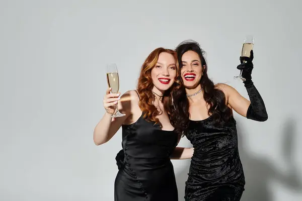Duas mulheres elegantes em vestidos pretos brindam alegremente com flautas de champanhe. — Fotografia de Stock