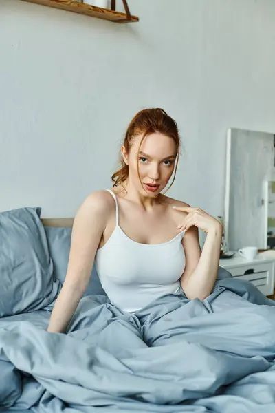 A woman in a white tank top sits atop a bed. — Stock Photo