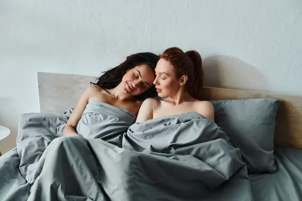 Due donne in elegante abbigliamento condividere un momento intimo sotto una coperta accogliente su un letto. — Foto stock