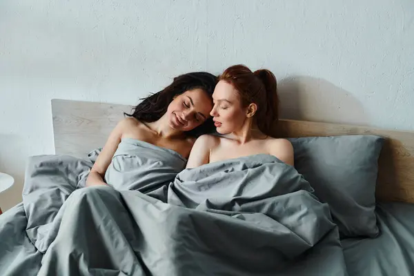Two women in elegant attire sharing an intimate moment under a cozy blanket on a bed. — Stock Photo