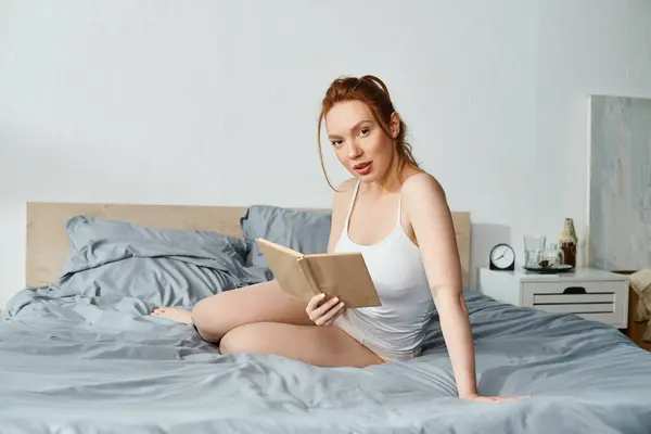A woman in elegant attire peacefully reads a book while sitting on a bed. — Stock Photo