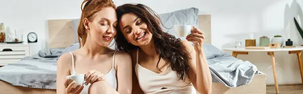 Loving lesbian couple in elegant attire, relaxing on bed, one woman holding a cup. — Stock Photo
