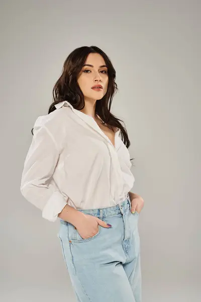 A beautiful plus size woman striking a pose in a white shirt and jeans against a gray backdrop. — Foto stock