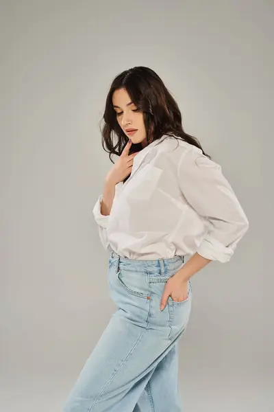 Красивая женщина с большим размером уверенно позирует в модной белой рубашке и синих джинсах на сером фоне. — стоковое фото