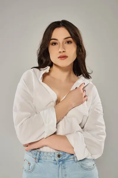 Stylish plus-size woman posing in trendy white shirt and jeans against a gray backdrop. — Stock Photo