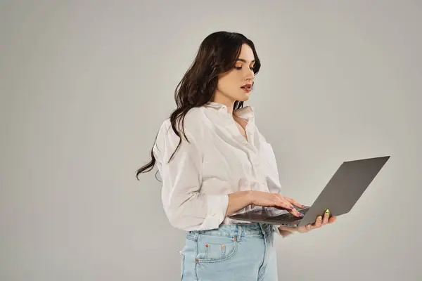 A beautiful plus size woman in stylish attire holds a laptop with confidence on a gray backdrop. — Stockfoto