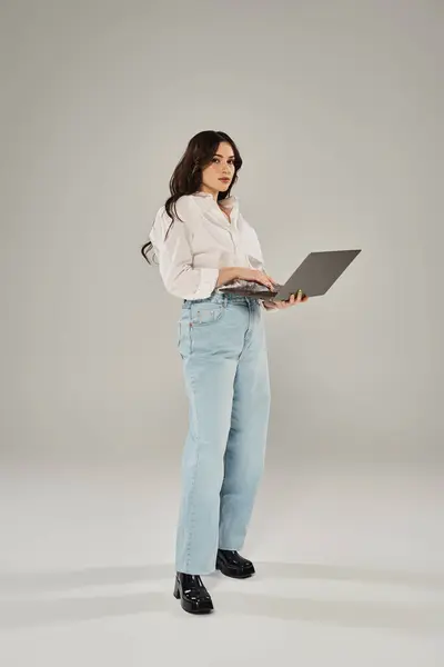 Плюс розмір жінка впевнено стоїть, тримаючи ноутбук у стильному вбранні на тлі сірого фону. — стокове фото