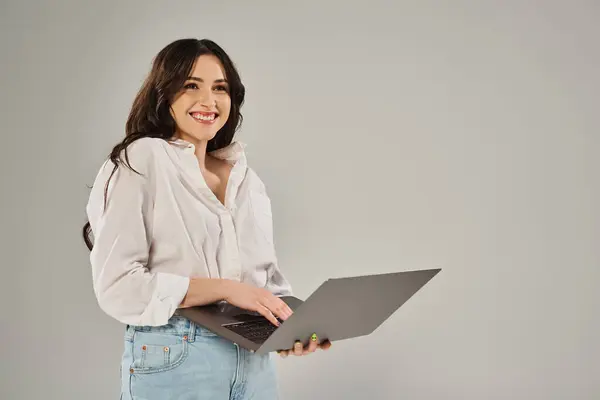 Eine stylische Plus-Size-Frau lächelt, während sie einen Laptop vor grauem Hintergrund hält. — Stockfoto