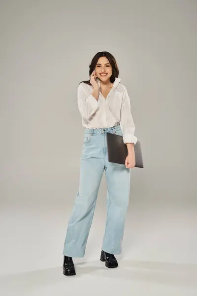 Femme taille plus en chemise blanche et jeans posant en toute confiance avec un ordinateur portable sur un fond gris. — Photo de stock