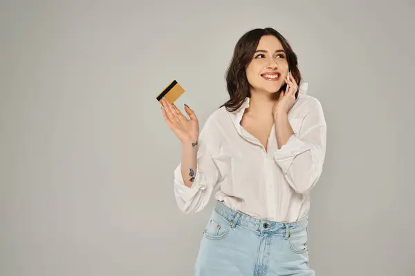Una mujer elegante de tamaño grande multitarea, sosteniendo una tarjeta de crédito y hablando en un teléfono celular contra un fondo gris. - foto de stock