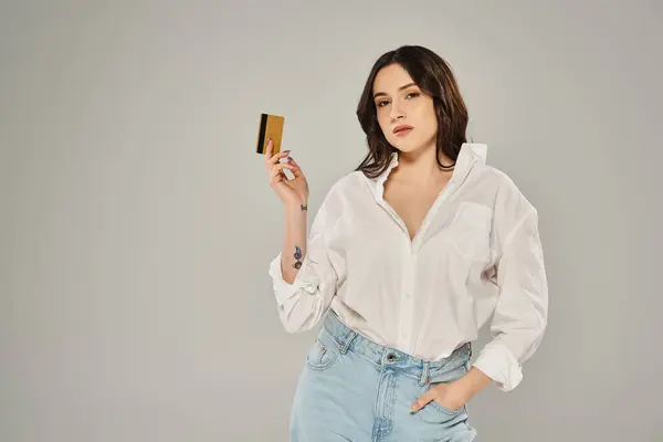 Beautiful plus-size woman in a white shirt confidently holding a gold card against a gray backdrop. — Stock Photo