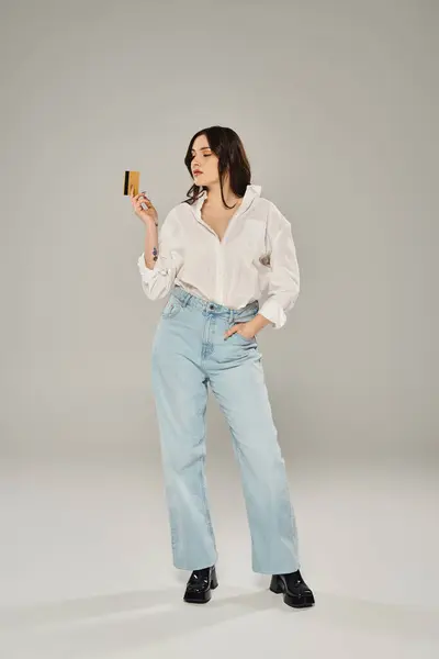 Mulher elegante plus-size em camisa branca delicadamente detém cartão de crédito contra um pano de fundo cinza elegante. — Fotografia de Stock
