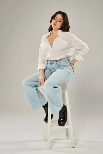 A stylish plus size woman in fashionable attire sitting elegantly on a white stool against a gray backdrop. — Stock Photo