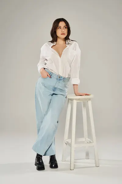 A beautiful plus size woman in a white shirt and jeans poses gracefully as she leans on a stool against a gray backdrop. — Stock Photo