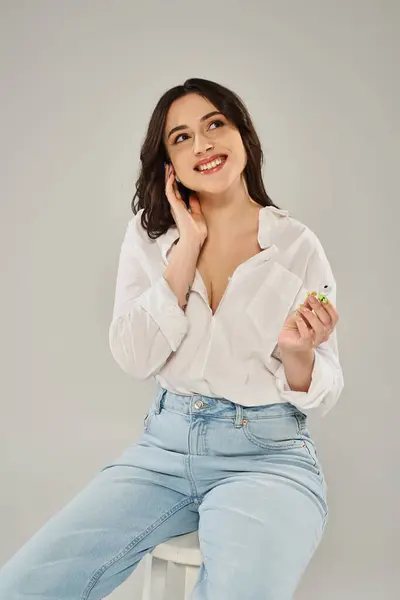 A beautiful plus size woman in stylish attire sits gracefully on a white stool against a gray backdrop. — Stock Photo