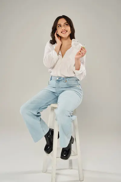 Une superbe femme de taille plus en tenue à la mode élégamment assise sur un tabouret blanc sur un fond gris. — Photo de stock