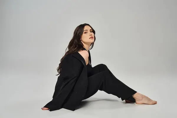 A beautiful plus-size woman in a stylish black suit sits gracefully on a grey backdrop, exuding confidence and poise. — Stock Photo