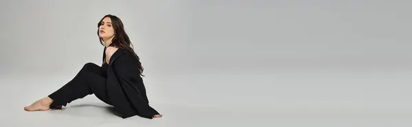 Uma bela mulher plus-size senta-se graciosamente no chão em uma roupa preta elegante contra um pano de fundo cinza. — Fotografia de Stock