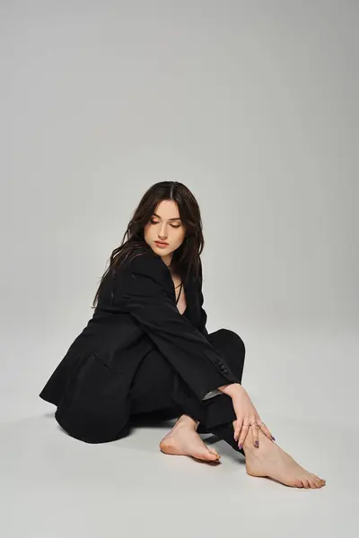 Una hermosa mujer de talla grande en un elegante abrigo negro sentado con gracia en el suelo contra un fondo gris. - foto de stock